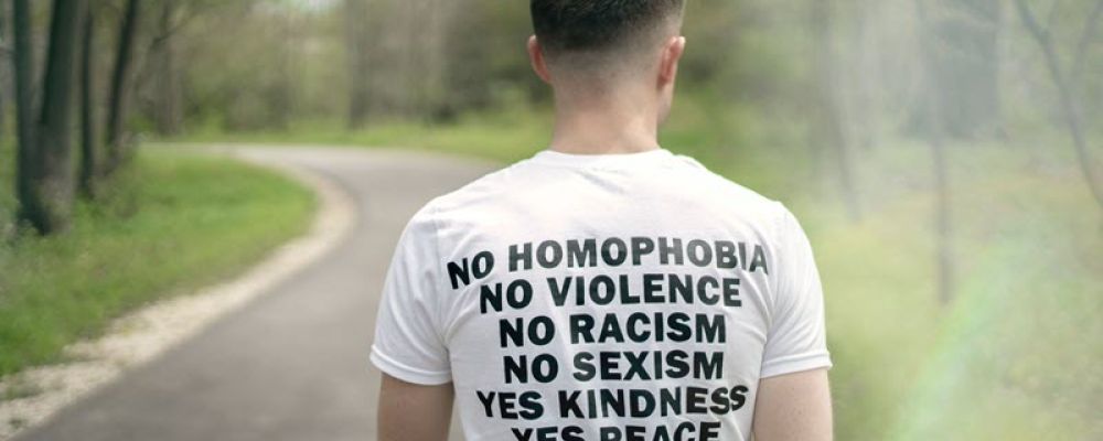 Person seen from behind walking down the road with a t-shirt on that has a list on back "No homophobia, no violence, no racism, no sexism, yes kindness, yes, peace, yes equality, yes love".